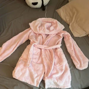 Rosa Morgonrock - Supermjuk och fluffig rosa morgonrock från Censored i storlek M/L. Jackan har huva med söta öron, knytskärp i midjan och stora fickor. Perfekt för mysiga dagar hemma eller när du vill vara extra cozy.