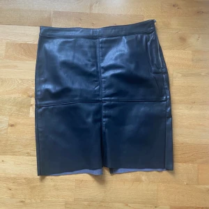 Svart skinnkjol från Vero Moda - Snygg svart skinnkjol från Vero Moda i storlek L. Kjolen är kort, har en markerad söm mitt fram och bak samt en slits bak för extra rörelsefrihet. Dragkedja i sidan ger en cool detalj. Perfekt för dig som gillar en edgy och stilren look.