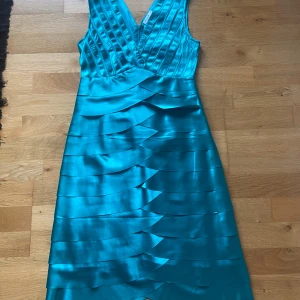 Turkos satin-klänning från Vila - Snygg turkos klänning från Vila i satin med omlott-v-ringning och breda axelband. Klänningen har ett lager-på-lager mönster och dragkedja. Helt oanvänd!