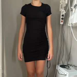 Svart kort klänning Divided H&M XS - Svart tight kortklänning från Divided H&M i storlek XS. Klänningen har korta ärmar, rund hals och snygg rynkdetalj med snörning på sidan. Mjuk och stretchig, perfekt för en trendig look.