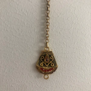 Unikt guldigt hängsmycke med röda stenar - Säljer ett hängsmycke med en cool, antikinspirerad berlock i guldton med svarta detaljer och röda stenar. Berlocken har en snygg form och fina snirkliga mönster. Perfekt för dig som vill sticka ut med något annorlunda och färgstarkt.