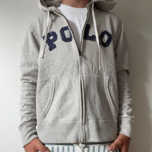 Grå hoodie från Polo Ralph Lauren - Snygg ljusgrå hoodie från Polo Ralph Lauren med blå POLO-tryck på bröstet. Tröjan har dragkedja, huva med snören och två stora fickor framtill. Perfekt för chill dagar och enkel att matcha med jeans eller joggers. Storlek L i barn men passar som S i herr. Modellen på bilden är 174 och väger ca 65kg.