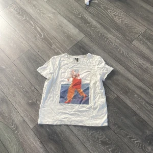 Vit t-shirt med print från Vero Moda - Vit t-shirt från Vero Moda med coolt tryck framtill som visar en person med orange byxor, röd väska och röda skor. Klassisk rund halsringning och korta ärmar. Skön och mjuk bomullskvalitet, perfekt till jeans eller kjol.