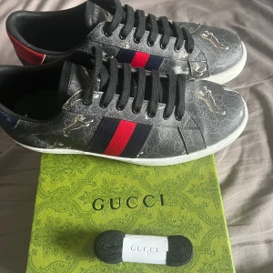 Gucci sneakers med tigrar och röd/blå rand - Snygga Gucci sneakers i mörkgrått med ikoniska röd/blå ränder på sidorna och coola tigermotiv. Skorna har svarta snören, vit sula och detaljer i rött på hälen. Kommer med originalkartong och extra skosnören. Perfekta för dig som vill sticka ut med lyxig streetstyle. Storlek 42 men passar 43