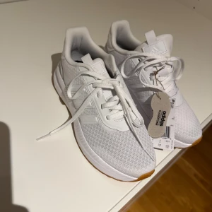 Vita adidas sneakers storlek 36 2/3 - Fräscha vita adidas sneakers med mesh-ovandel och klassiska tre ränder på sidan. Skorna har snörning och en platt sula med gummidetalj framtill. Perfekta för dig som gillar sportig och clean stil.