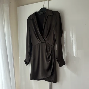 Mörkbrun omlottklänning från Zara - Snygg mörkbrun kort omlottklänning från Zara i satinliknande material. Klänningen har v-ringning, långa ärmar med manschett och krage. Sitter figurnära och har en draperad detalj i midjan för extra stilpoäng.