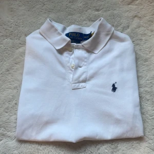 Vit pikétröja från Ralph Lauren - Klassisk vit pikétröja från Ralph Lauren med blå broderad logga på bröstet. Tröjan har korta ärmar, krage och knäppning med två knappar framtill. Tillverkad i mjuk bomull som känns skön mot huden. Perfekt för en clean och stilren look.