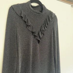 Glittrig svart långärmad topp med volang - Supersnygg svart glittrig topp med lång ärm och volangdetalj framtill. Hög hals och ett lätt stretchigt material som sitter bekvämt. Perfekt för dig som vill sticka ut lite extra med en cool och trendig look.