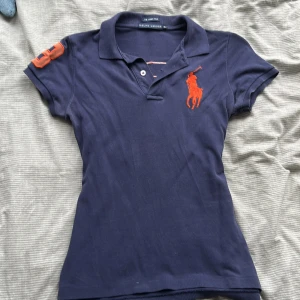Marinblå pikétröja Ralph Lauren XS - Snygg marinblå pikétröja från Ralph Lauren i modellen The Skinny Polo. Tröjan har orange logga på bröstet, stor orange 3:a på ryggen och ärm, samt klassisk krage och korta ärmar. Perfekt för en sportig och stilren look.