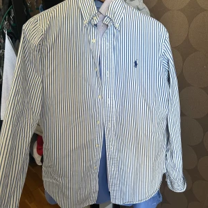 Randig skjorta från Polo Ralph Lauren - Säljer en klassisk vit skjorta med blå ränder från Polo Ralph Lauren. Skjortan har lång ärm, knappar framtill och den ikoniska broderade loggan på bröstet. Modellen är custom fit och passar perfekt till dig som gillar en stilren look.