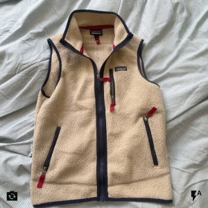  fleeceväst patagonia - Säljer en beige fleeceväst från Patagonia i storlek S. Västen har marinblå detaljer, hel dragkedja framtill och tre fickor med röda zippar. Perfekt för lager-på-lager och har en hög krage för extra värme. Klassisk och stilren design.