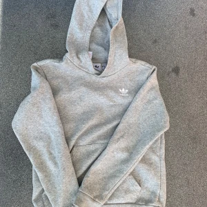 Grå hoodie från Adidas Originals - Säljer en stilren grå hoodie från Adidas Originals med klassisk vit logga på bröstet. Tröjan har huva, magficka och långa ärmar. Perfekt för chill dagar eller när du vill ha en enkel och snygg look.