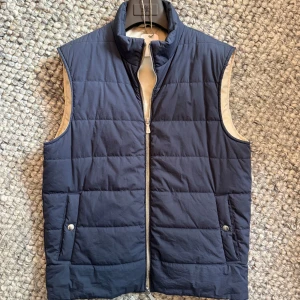 Eleventy vest - Snygg mörkblå dunväst från Eleventy med quiltad design och dragkedja framtill. Västen har två fickor med knappstängning och en beige insida som ger en clean kontrast. Perfekt för lager-på-lager och stilren look.
