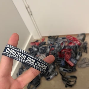 Christian Dior J'Adior armband blå - Snyggt vävt armband från Christian Dior med texten 'J'Adior' i vitt på marinblå bakgrund. Armbandet har justerbar knytning med tofsar i ändarna och coola detaljer längs kanterna. Perfekt accessoar för att lyfta din outfit med en lyxig touch.