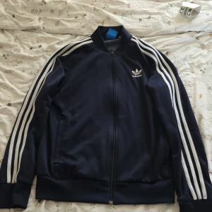 Adidas blå tröja med ränder - Klassisk marinblå Adidas tröja med vita ränder längs ärmarna och dragkedja framtill. Tröjan har ribbade muddar och den ikoniska Adidas-loggan på bröstet. Perfekt för en sportig och avslappnad stil.