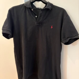Svart pikétröja från Polo Ralph Lauren - Klassisk svart pikétröja från Polo Ralph Lauren med röd broderad logga på bröstet. Tröjan har korta ärmar, krage och knäppning med två knappar. Tillverkad i bomull för en skön och avslappnad känsla. Perfekt för en stilren och sportig look.
