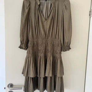 Olivgrön kortklänning från Maje - Supersöt olivgrön kortklänning från Maje med volanger, v-ringning och knytband vid halsen. Klänningen har långa ärmar med smock och volangdetaljer vid ärmslut samt flera lager i kjolen för en flowy look. Perfekt för dig som gillar en avslappnad men chic stil. Använd 1 gång 
