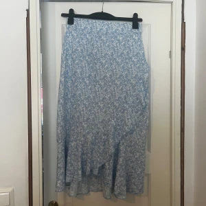 Blå blommig sommarkjol - Supersöt långkjol i ljusblått med vitt blommönster. Kjolen har omlottmodell och volangkant nertill. Endast använd en gång