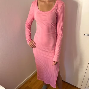 Rosa ribbad långklänning med slits - Snygg rosa långklänning med ribbad struktur och lång ärm. Klänningen har en rundad urringning och en slits på sidan som ger en extra touch. Figurnära passform och stretchigt material gör den både bekväm och trendig.