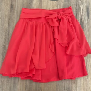 Röd omlottkjol med volang och knyt - Superfin röd kortkjol i omlottmodell med volangdetaljer och bred knytrosett i midjan. Kjolen har en lätt och flowy känsla, perfekt för att sticka ut. Stängs med dragkedja i sidan och har en feminin look.