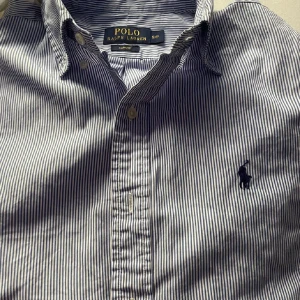 Randig blåvit skjorta Polo Ralph Lauren - Snygg blåvit randig skjorta från Polo Ralph Lauren i slim fit. Klassisk krage, långa ärmar och knappar framtill. Broderad logga på bröstet. Perfekt till jeans eller chinos för en clean och stilren look.