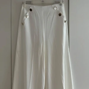 Vita vida byxor med knappar - Snygga vita byxor med hög midja och vida ben. Byxorna har dekorativa guldiga knappar framtill och är sydda i ett mjukt, vävt tyg som ger en clean och stilren look. Perfekta för dig som gillar minimalistisk och trendig stil