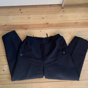 Svarta byxor med dragsko från New Fashion - Svarta byxor från New Fashion med elastisk midja och dragsko. Byxorna har raka ben, dragkedjefickor och är tillverkade i ett mjukt tyg. Insidan har tunna vita ränder. Perfekta för en chill och stilren look.