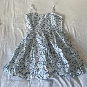 Vit och blå blommig sommarklänning H&M - Superfin kort klänning från H&M med smala axelband och blått blommönster på vit botten. Klänningen har smockad rygg och en lätt utställd kjol, perfekt för varma dagar. Tillverkad i mjuk bomull som känns skön mot huden.