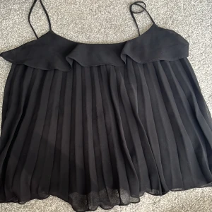 Svart plisserad linnetopp med volang - Supersöt svart linnetopp med tunna axelband och volangdetalj upptill. 