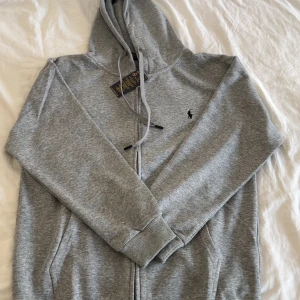 Ralph lauren zip - PRIS ÄR INTE HUGGET I STEN!🤩👌                 Färg: Ljusgrå 	•	Storlek: M 	•	Skick: Helt ny, aldrig använd 	•	Detaljer: Hoodie med dragkedja framtill, fickor på sidorna och svart Polo Ralph Lauren-logga broderad på bröstet. Justerbar huva med dragsko. 	•	Material: Mjuk bomullsblandning som passar både sommar- och höstväder. 