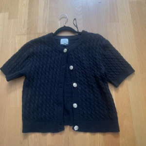 Svart stickad kofta med guldknappar - Kortärmad svart stickad kofta med snyggt kabelstickat mönster och dekorativa guldiga knappar framtill. 