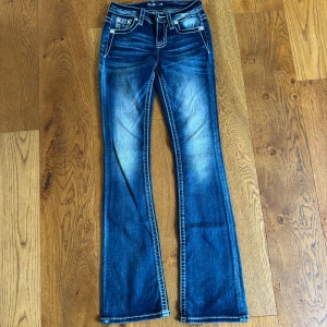 Miss Me blå bootcut jeans 24/34 - Snygga blå bootcut jeans från Miss Me med kontrastsömmar och broderade bakfickor i turkos, beige och svart. Jeansen har låg midja, klassisk femficksdesign och coola detaljer på knappar och nitar. Perfekta för dig som gillar statement-jeans!