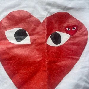 Vit Comme des Garçons Play t-shirt - Säljer en vit t-shirt från Comme des Garçons Play med röda hjärtat med ögon tryckt på bröstet. T-shirten är i bomull och har en klassisk passform med korta ärmar. 