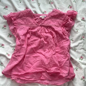 Rosa blus från Gina Tricot - Supersöt rosa blus från Gina Tricot med volangärmar och v-ringning. Blusen har smockdetaljer upptill och är tillverkad i ett lätt och luftigt tyg.