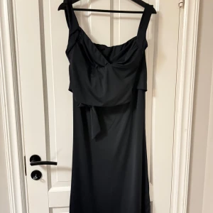 Svart set med lång kjol och en rygglös top med knyt - Stilren svart långklänning med breda axelband och snygg knytning framtill. Klänningen har en elegant, enkel design och är perfekt för dig som gillar en klassisk look. Materialet känns mjukt och följsamt mot kroppen.