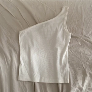Vit oneshoulder topp från Kappahl - Säljer en stilren vit oneshoulder topp från Kappahl. Toppen är ribbad och har en ärmlös design med en axelrem, vilket ger en snygg och modern look. Perfekt att matcha med jeans eller kjol för en trendig outfit.