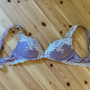 Lila spets-bh med blommig detalj - Superfin lila bh med vackra blommiga spetsdetaljer i vitt. Kuporna är lätt vadderade och har en elegant spetskant runt om. Axelbanden är justerbara och bh:n har en mjuk känsla mot huden. Perfekt för dig som gillar romantiska och feminina underkläder.