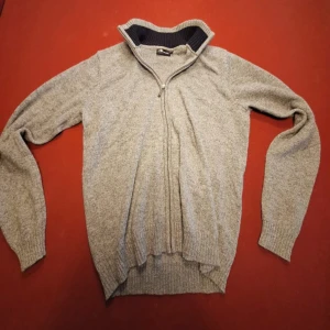 Grå stickad tröja Camargue half zip - Mysig grå stickad tröja från Camargue med ribbad krage i mörkblått och grått. Tröjan har half zip-dragkedja och långa ärmar, perfekt för kyliga dagar. Enkel och stilren design som funkar till det mesta. Storlek M men passar mer för S