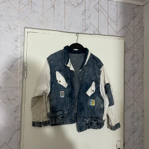Retro jeansjacka med vita ärmar, stl 36 - Säljer en unik jeansjacka i retrostil med blå denim och kontrasterande vita ärmar. Jackan har två stora fickor framtill med tryckknappar och coola patches som detaljer. Perfekt för dig som vill sticka ut med en streetig vibe.