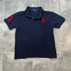 Marinblå pikétröja från Polo Ralph Lauren - Klassisk marinblå pikétröja från Polo Ralph Lauren med röd broderad logga på bröstet och rött nummer 3 på ärmen. Tröjan passar dig som är runt 170. Liter hål på magen men märks knappt och kan enkelt sys igen. MVH LUNDGRENS