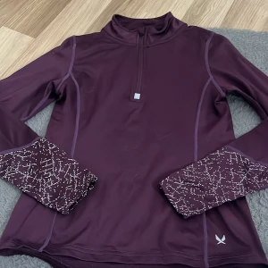Vinröd träningströja SOC half zip - Snygg vinröd långärmad träningströja från SOC med half zip och coola vita prickdetaljer på ärmsluten. Tröjan är i syntetmaterial som andas och har en sportig passform, perfekt för aktiva dagar. Stilsäkra sömmar och diskret logga framtill.