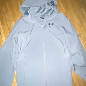 Grå vindjacka från Under Armour - Snygg grå vindjacka från Under Armour med huva och dragkedja framtill. Jackan har logga på bröstet och diskret text längs dragkedjan. Perfekt för dig som gillar sportig stil och vill ha något lätt och smidigt till vardagen. Har använts 3-4 ggr 