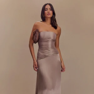 Champagnefärgad axelbandslös långklänning - Snygg axelbandslös långklänning i champagnefärgad satin-look. Klänningen har figurnära passform, snörning i ryggen och lätt rynkade detaljer vid midjan. Perfekt för dig som vill ha en elegant och stilren look med en touch av lyx. Klänningen är oanvänd 