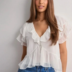 Vit volangblus med v-ringning - Supersöt vit blus i transparent material med volanger längs ärmar och nederkant. Djup v-ringning framtill och korta, luftiga ärmar ger en romantisk vibe. Perfekt att styla med jeans eller kjol för en trendig look.