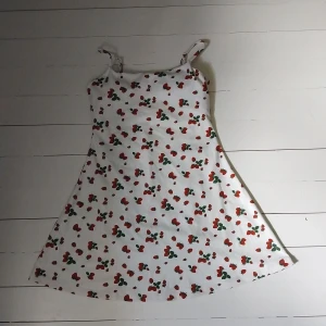 Vit kortklänning med jordgubbsmönster - Supersöt vit kortklänning med röda jordgubbar och gröna blad som mönster. Klänningen har smala axelband och en lätt utställd passform. Den har även inbygda shorts med fickor likt en 'skort', fast som klänning, och även en inbyggd topp med pads. Aldrig använd - etikett sitter kvar.