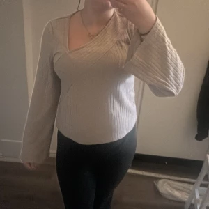 Beige omlottstickad tröja - Säljer en beige stickad tröja med omlottdesign och v-ringning. Tröjan har långa ärmar och ribbad struktur som ger en snygg och modern look. Perfekt för dig som gillar stilrena och bekväma plagg med det lilla extra.