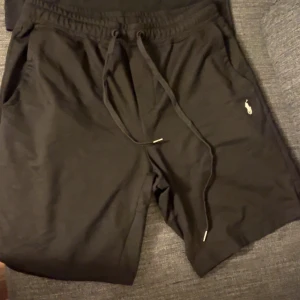 Svarta shorts från Polo Ralph Lauren - Svarta shorts från Polo Ralph Lauren med klassisk vit logga broderad på vänster ben. De har elastisk midja med snörning och två sidofickor. Perfekta för chill dagar eller när du vill ha en clean och enkel look.