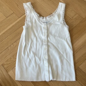 Vit spetskantad linnetopp Brandy Melville - Supersöt vit ribbad linnetopp från Brandy Melville med spetskant runt halsringning och ärmhål. Knappar hela vägen framtill ger en gullig touch. Perfekt för lager-på-lager eller sommarlooken. Man kan öppna alla knappa
