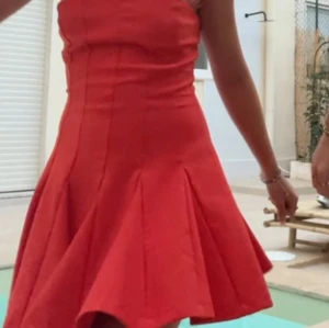 Orange axelbandslös kortklänning - Säljer en röd axelbandslös klänning med utsvängd kjol och markerad midja. Klänningen har en figurnära överdel och flera sömmar som ger en snygg passform. Perfekt för dig som vill sticka ut med en färgstark look.