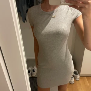Grå ribbad kortklänning - Säljer en tight, ribbad grå kortklänning med korta ärmar och rund hals. Klänningen är enkel och stilren, perfekt för dig som gillar en clean look. Materialet är mjukt och stretchigt för en bekväm passform.